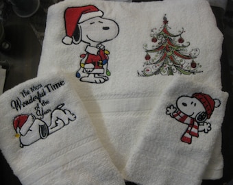May include: Set of four white towels with embroidered Christmas designs. One towel features a red-hatted Snoopy with the text "The Most Wonderful Time of the Year". Another towel features a red-hatted Snoopy with Christmas lights. A third towel features a Christmas tree with ornaments. The fourth towel features a red-hatted Snoopy with a red and white scarf.