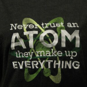 May include: A black t-shirt with a white embroidered design. The design features a stylized atom with the text "Never trust an ATOM they make up EVERYTHING".