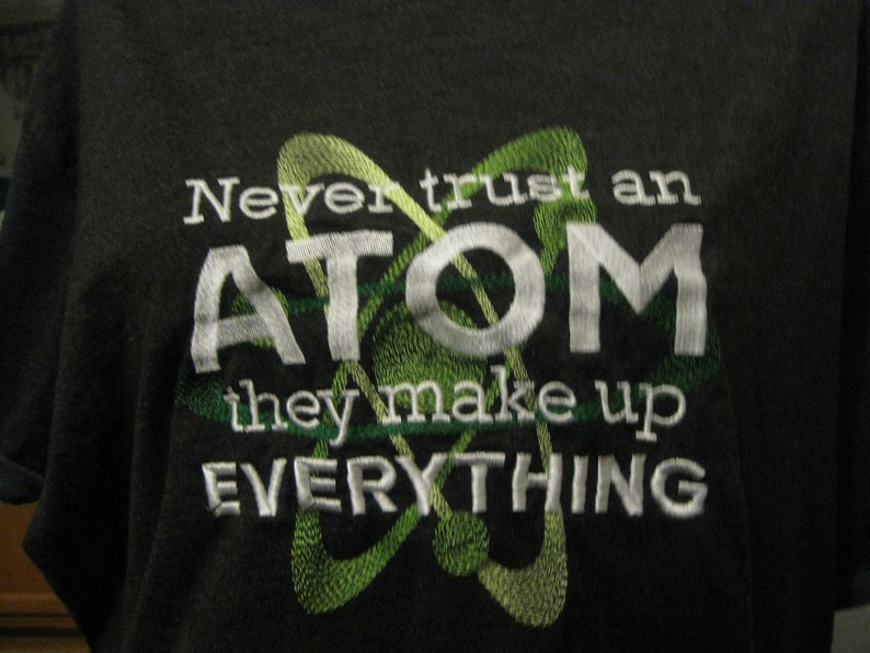 May include: A black t-shirt with a white embroidered design. The design features a stylized atom with the text "Never trust an ATOM they make up EVERYTHING".