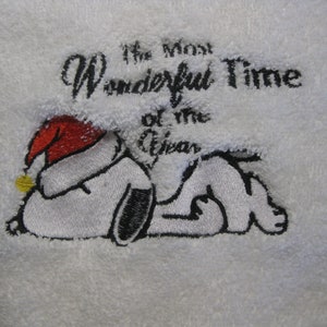 Op de afbeelding: Witte geborduurde handdoek met een zwart-witte cartoon hond die een rode kerstmanmuts draagt. De tekst "The Most Wonderful Time of the Year" is geborduurd boven de hond.