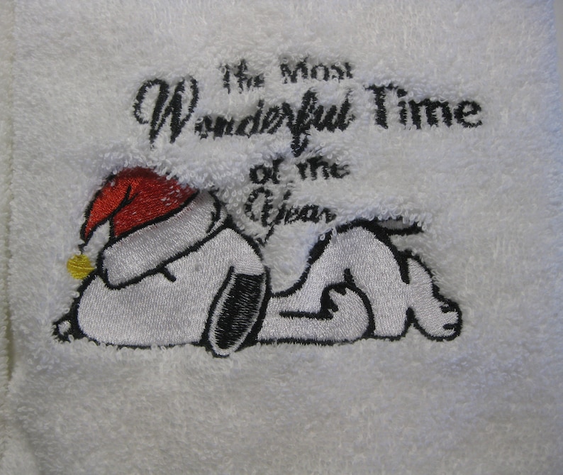 Op de afbeelding: Witte geborduurde handdoek met een zwart-witte cartoon hond die een rode kerstmanmuts draagt. De tekst "The Most Wonderful Time of the Year" is geborduurd boven de hond.