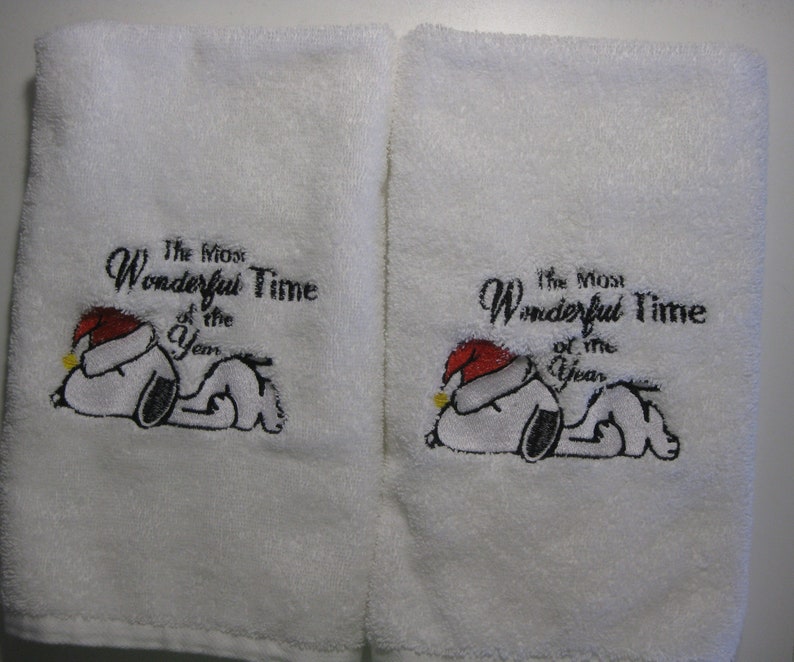 Op de afbeelding: Twee witte handdoeken met geborduurde afbeeldingen van Snoopy met een kerstmanmuts. De tekst "The Most Wonderful Time of the Year" is op elke handdoek geborduurd.