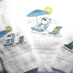 May include: White bath towels with embroidered beach scene featuring a cartoon dog and bird relaxing in beach chairs under a blue umbrella and sun. The towels have a blue border.
