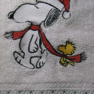 May include: Embroidered white towel with a red and white Snoopy wearing a Santa hat and a red scarf.  Snoopy is walking with Woodstock, who is also wearing a red scarf.  The towel has a green embroidered border.