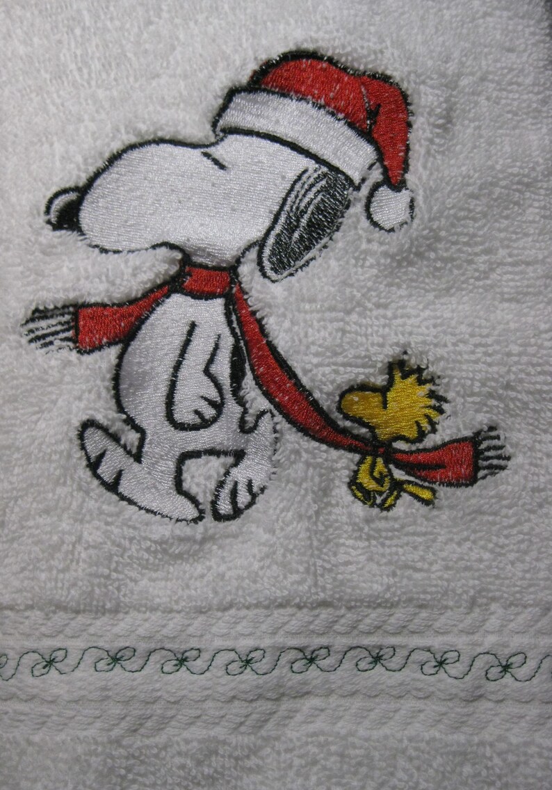May include: Embroidered white towel with a red and white Snoopy wearing a Santa hat and a red scarf.  Snoopy is walking with Woodstock, who is also wearing a red scarf.  The towel has a green embroidered border.
