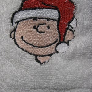 May include: Embroidered white towel with a cartoon character wearing a red Santa hat. The character has a black outline and a smiling face.