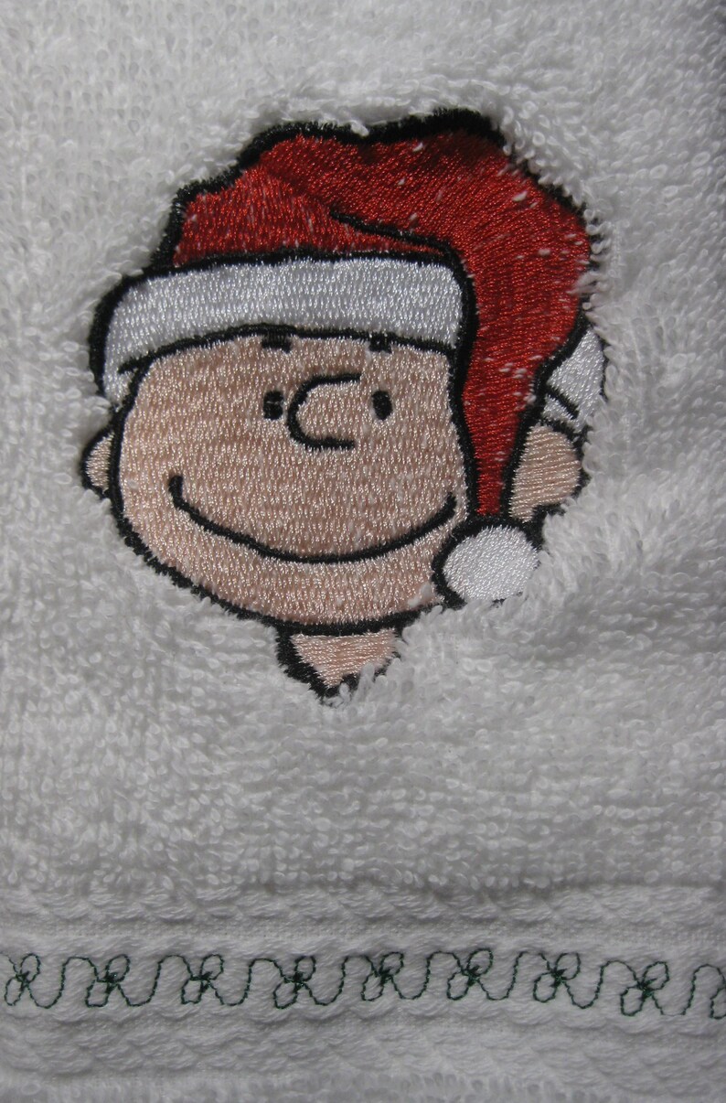 May include: Embroidered white towel with a cartoon character wearing a red Santa hat. The character has a black outline and a smiling face.