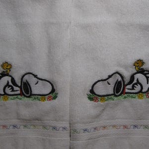 May include: Two white towels with embroidered images of Snoopy and Woodstock. Snoopy is lying down with his eyes closed and Woodstock is perched on his back. Both images are surrounded by small embroidered flowers.