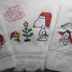 May include: Three white towels with embroidered images of Snoopy and Woodstock. The towels feature a Christmas theme with Snoopy and Woodstock wearing Santa hats. The text "May your Days be Merry and Bright" is embroidered on one of the towels.