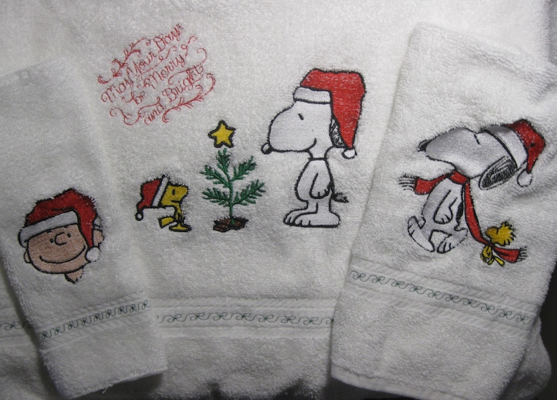 May include: Three white towels with embroidered images of Snoopy and Woodstock. The towels feature a Christmas theme with Snoopy and Woodstock wearing Santa hats. The text "May your Days be Merry and Bright" is embroidered on one of the towels.