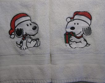 May include: Two white bath towels with embroidered images of Snoopy wearing a Santa hat. One Snoopy is holding a gift box and the other is holding a candy cane.