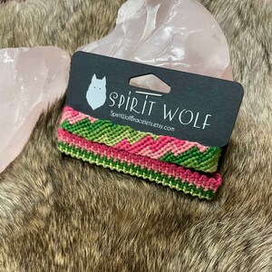 May include: Two green and pink friendship bracelets with a chevron pattern. The bracelets are on a black card with the text "Spirit Wolf" and "SpiritWolfBracelets.etsy.com".