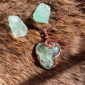 May include: A green fluorite crystal pendant wrapped in copper wire, hanging from a copper chain. The pendant is surrounded by two smaller fluorite crystals on a brown fur background.