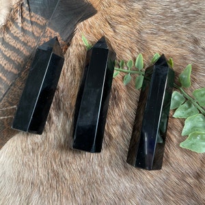 May include: Three polished black obsidian obelisks with pointed tops. The obelisks are displayed on a textured, brown surface with a feather and greenery in the background. The obelisks are a dark, glossy black.