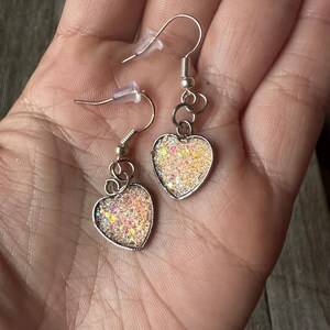 May include: Silver-toned heart-shaped earrings with iridescent glitter filling. The earrings have a hook closure and are held in a hand. The heart-shaped pendants are outlined in silver.