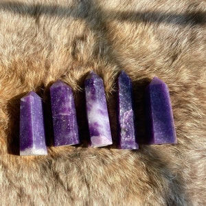 May include: Five purple amethyst crystal points arranged in a row on a brown faux fur background.
