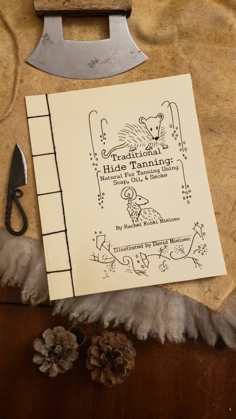 Op de afbeelding: Een boekomslag met een beige achtergrond en zwarte illustraties van een stekelvarken, een ram en een bloemmotief. De titel is "Traditional Hide Tanning: Natural Fur Tanning Using Soap, Oil, & Smoke". Het boek is van Rachel Koski Nielsen en ge&iuml;llustreerd door David Nielsen.