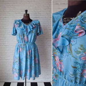 May include: A vintage light blue dress with a floral pattern. The dress features short sleeves, a ruffled neckline, and a cinched waist. The floral design includes pink, green, and white accents. The dress is displayed on a mannequin.