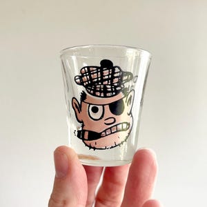 May include: A clear glass shot glass featuring a printed cartoon illustration of a person wearing a plaid tam o' shanter hat, an eye patch, and holding a cigar in their mouth. The design is rendered in black, white, and light pink tones.