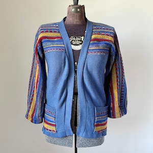 Peut inclure: Un cardigan bleu clair avec des accents rayés colorés sur les manches et les poches. Le pull a un col en V et deux poches avant. Les manches et les épaules supérieures présentent des motifs rayés rouges, jaunes et bleus. Le pull est présenté sur un mannequin.