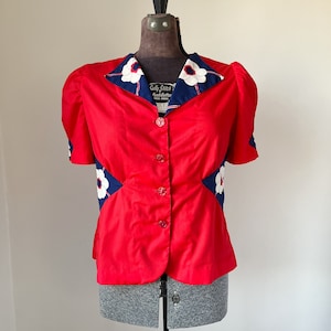 Puede incluir: Una blusa roja vintage con botones y mangas cortas. El cuello y los detalles de las mangas presentan una tela azul marino con estampados florales blancos. La blusa se exhibe en un maniquí.