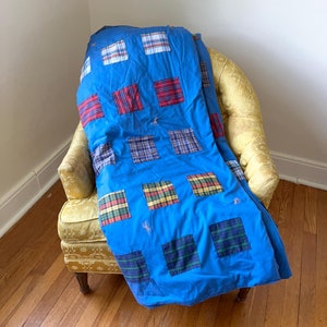 May include: A blue patchwork quilt with plaid squares in red, green, and blue covers a yellow armchair. The quilt has a solid blue background and is draped over the chair, showcasing the colorful plaid patterns.