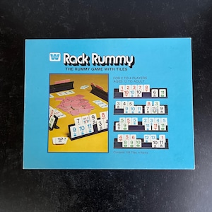 May include: A Rack Rummy game box with a light blue background. The box features the game's title and a graphic showing game tiles and racks. The text on the box reads "FOR 2 TO 4 PLAYERS AGES 12 TO ADULT".