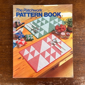 May include: The cover of "The Patchwork Pattern Book" is displayed, showcasing a red and white patterned placemat with a fork and spoon. A green and white placemat is also visible, along with small decorative pillows and a bowl of strawberries.