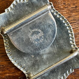 May include: A silver-toned metal belt buckle with a scalloped edge and a rectangular frame. The back features a circular stamp with the text CRAFTED WITH PRIDE WAGES IN U.S.A. and includes a hinged bar and a hook for securing the belt strap.