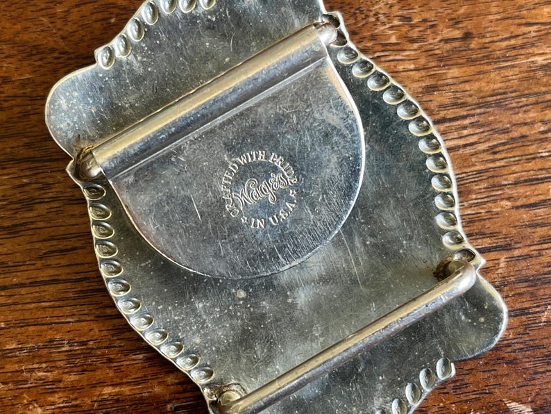 May include: A silver-toned metal belt buckle with a scalloped edge and a rectangular frame. The back features a circular stamp with the text CRAFTED WITH PRIDE WAGES IN U.S.A. and includes a hinged bar and a hook for securing the belt strap.