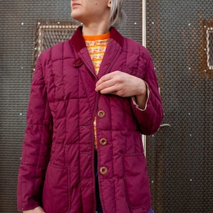 May include: A quilted burgundy jacket with a corduroy collar and three large brown buttons. The jacket has two front pockets and a tailored fit. The person is wearing a patterned sweater underneath and tie-dye pants.