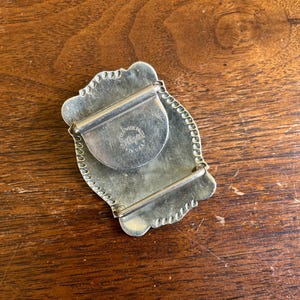 May include: A silver-toned metal belt buckle with a scalloped edge and a rectangular frame. The back features a central circular plate stamped with the text "SOLID BRASS" and "MADE IN U.S.A." along with a central brand logo mark.