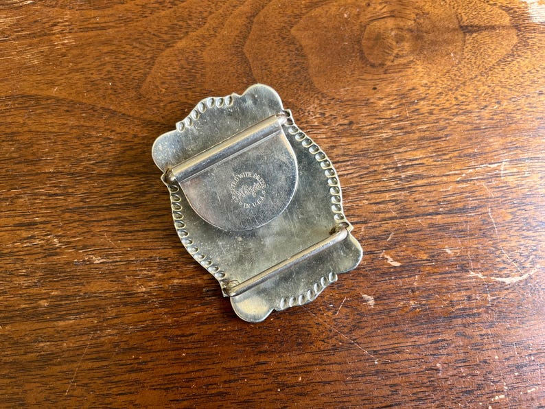 May include: A silver-toned metal belt buckle with a scalloped edge and a rectangular frame. The back features a central circular plate stamped with the text "SOLID BRASS" and "MADE IN U.S.A." along with a central brand logo mark.