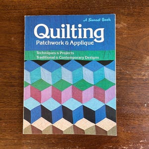 May include: A Sunset Book titled "Quilting Patchwork & Applique" with a blue cover and geometric design. The cover features the title in white, with text below indicating techniques, projects, and designs. The background is a wooden surface.