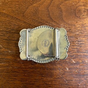 May include: A rectangular brass belt buckle with scalloped edges and a beaded border. The back features a hinged metal bar and a circular stamp that reads "Bergamot Brass Works" and "Made in U.S.A." centered on the mechanism.