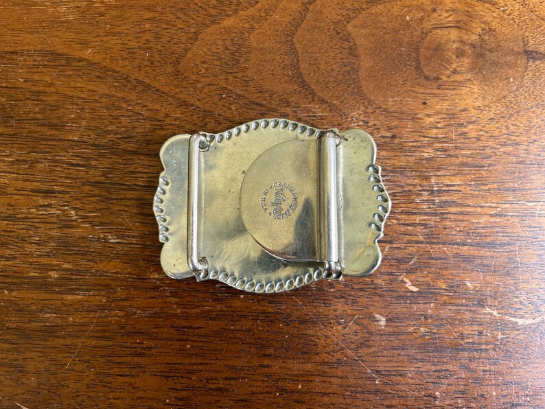 May include: A rectangular brass belt buckle with scalloped edges and a beaded border. The back features a hinged metal bar and a circular stamp that reads "Bergamot Brass Works" and "Made in U.S.A." centered on the mechanism.