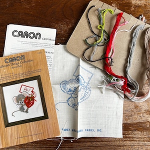 May include: A vintage Hallmark Design Collection crewel kit featuring a framed mouse holding a heart. The kit includes embroidery floss in various colors, a printed fabric, and instructions. The kit is on a wooden surface.