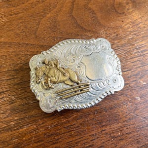 May include: A rectangular silver-tone belt buckle featuring an embossed floral border and a rope-style edge. A brass-colored relief depicts a rider on a horse jumping over a fence. The buckle includes a smooth, blank shield-shaped area for engraving.