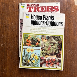 May include: Two vintage gardening books, "The World of Trees" and "House Plants Indoors/Outdoors," are stacked on a wooden surface. The "House Plants" book features images of plants and flowers. The books are from "Ortho Books."