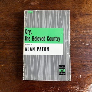 May include: A vintage paperback book titled "Cry, the Beloved Country" by Alan Paton. The book has a gray cover with vertical lines and a green and white title block. The Scribner Library logo is visible.