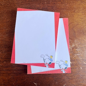 May include: Two sheets of white stationery paper with a red brick pattern border. Each sheet features a graphic of Snoopy wearing a blue cap and holding a paint roller, with Woodstock perched on top. The text reads Snoopy 1958 United Feature Syndicate, Inc.