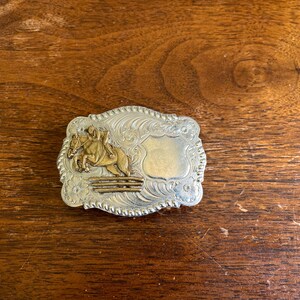 May include: A rectangular silver-toned belt buckle with a rope-style border and intricate scrollwork engraving. The center features a raised gold-toned figure of a horse and rider jumping over a fence, alongside a smooth, blank shield-shaped area.