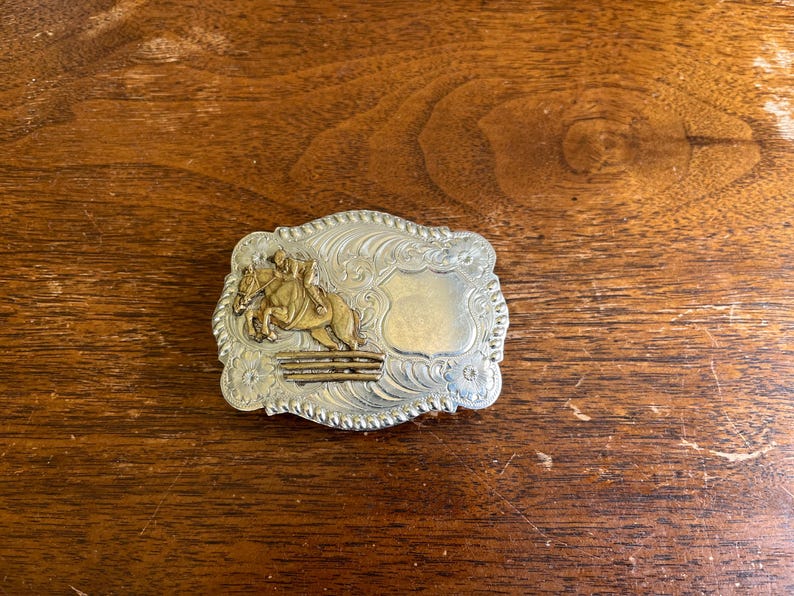 May include: A rectangular silver-toned belt buckle with a rope-style border and intricate scrollwork engraving. The center features a raised gold-toned figure of a horse and rider jumping over a fence, alongside a smooth, blank shield-shaped area.