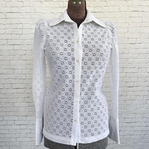 May include: White eyelet shirt with a collared neckline and long sleeves. The shirt features a floral pattern and button-down front. The sleeves have a flared cuff. The shirt is displayed on a mannequin against a white brick wall.