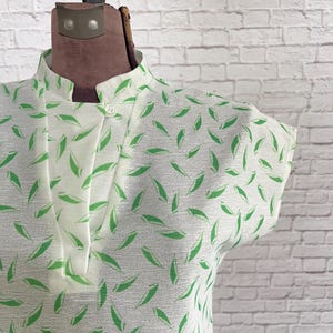 Puede incluir: Una blusa blanca de manga corta con un patrón repetitivo de hojas verdes, cuello mao y un cierre de un solo botón blanco en el escote. La prenda se muestra sobre un maniquí, destacando una tela texturizada y un corte estructurado.