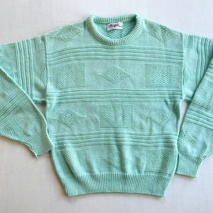 Peut inclure: Un pull vert menthe avec un col rond et des manches longues. Le pull présente un motif texturé de rayures horizontales, de carrés et de losanges. Les poignets et l'ourlet sont côtelés.