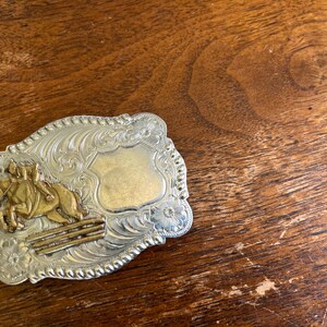 May include: A rectangular silver-tone belt buckle with a rope-style border and floral engravings. The buckle features a gold-tone relief of a horse and rider jumping over a fence on the left side and a smooth, blank shield-shaped area on the right.