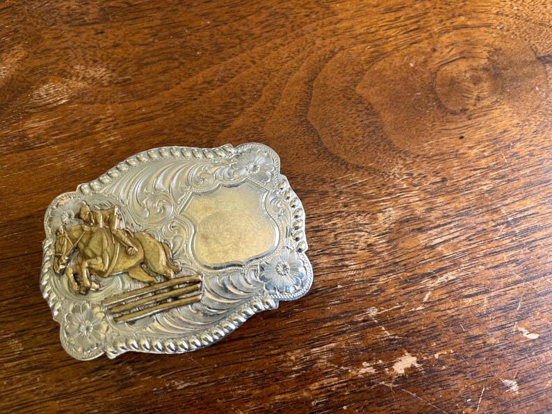 May include: A rectangular silver-tone belt buckle with a rope-style border and floral engravings. The buckle features a gold-tone relief of a horse and rider jumping over a fence on the left side and a smooth, blank shield-shaped area on the right.