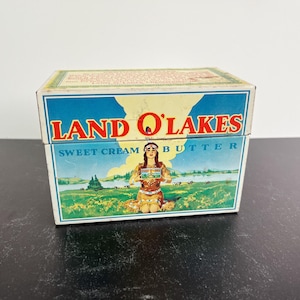 Puede incluir: Una caja de mantequilla Land O'Lakes Sweet Cream vintage. La caja presenta un diseño azul y amarillo con el nombre de la marca en rojo. El panel frontal representa a una mujer nativa americana en un campo con un lago y árboles. La caja es rectangular.