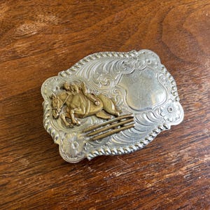 May include: A rectangular silver-tone belt buckle with a rope-style border and intricate floral scrollwork. The center features a raised brass-colored relief of a horse and rider jumping over a fence, alongside a smooth, blank shield-shaped area.